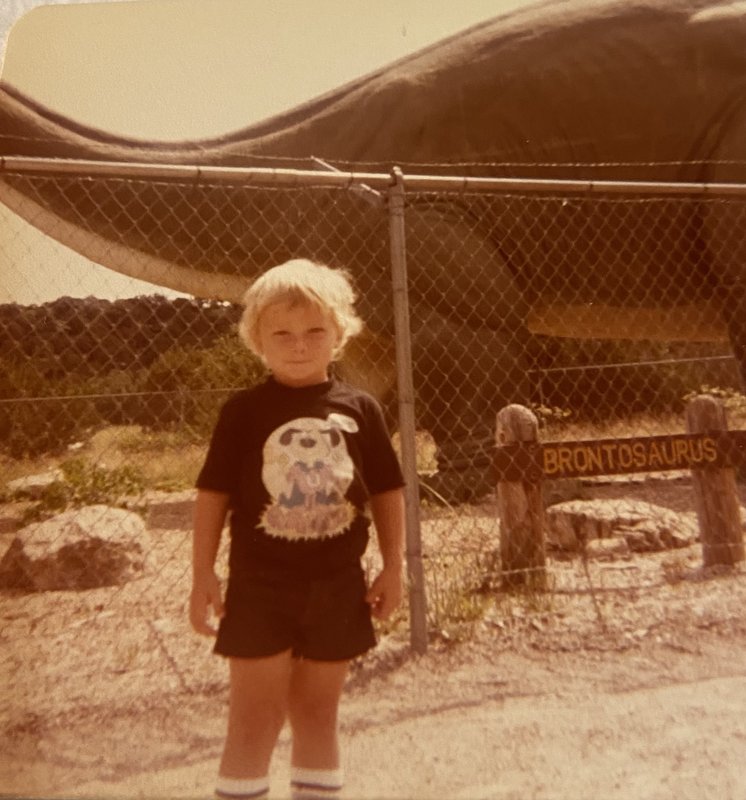 Young Jessie Heath — a blonde boy standing in front of a Brontosaurus sign